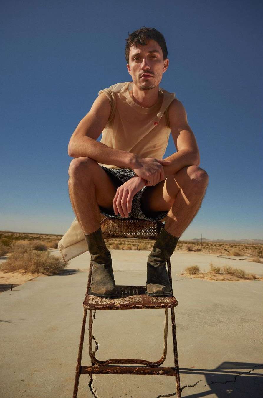 A fair-skinned adult male sits on an old metal chair outdoors in a desert landscape wearing casual clothes on a sunny day.