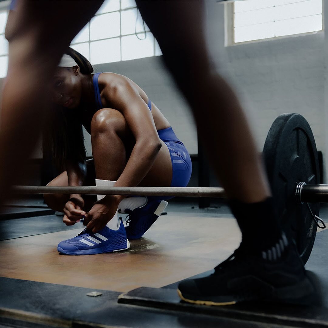 A woman in athletic wear tying her sneaker while squatting in a gym with a barbell, showing fitness and dedication in an indoor setting.