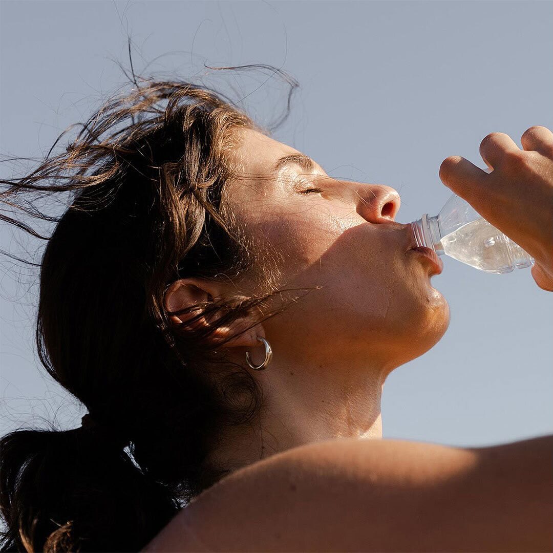 Woman drinks water from a transparent plastic bottle outdoors, close-up shot, bright natural lighting, blue sky. Hydration and health.