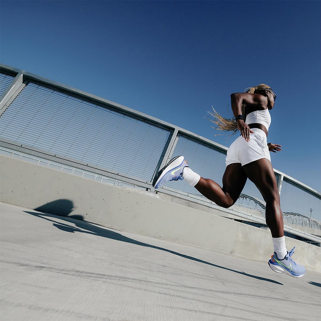 Athletic African-American woman sprints across a modern urban bridge in white athletic clothing and light blue running shoes on a sunny day.