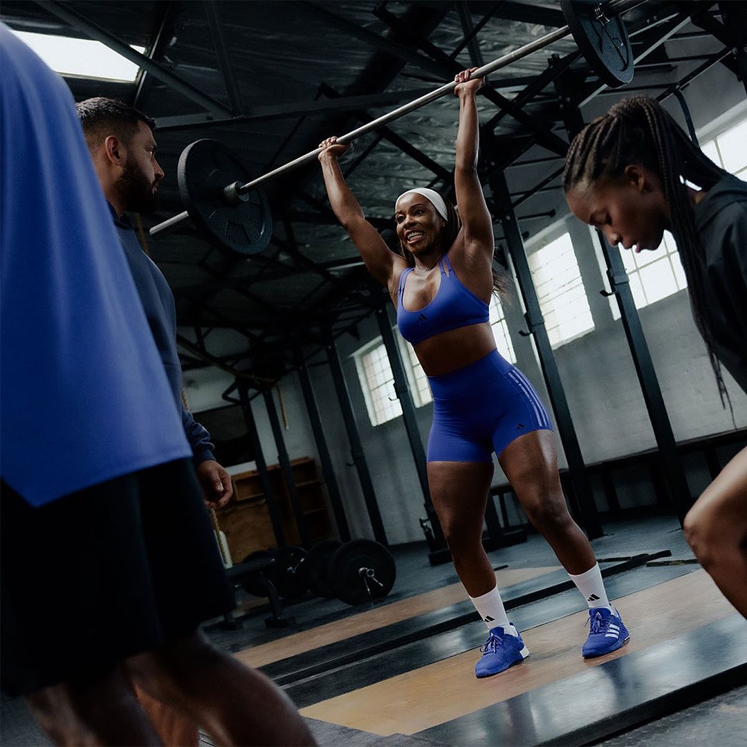 A woman in a blue sports bra and shorts is lifting a barbell overhead, surrounded by other people in a gym with a concrete floor.