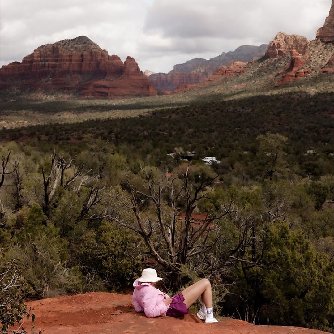 Woman rests on red rock amidst a desert landscape, wearing a pink jacket, purple shorts, white shoes and a light-colored hat.