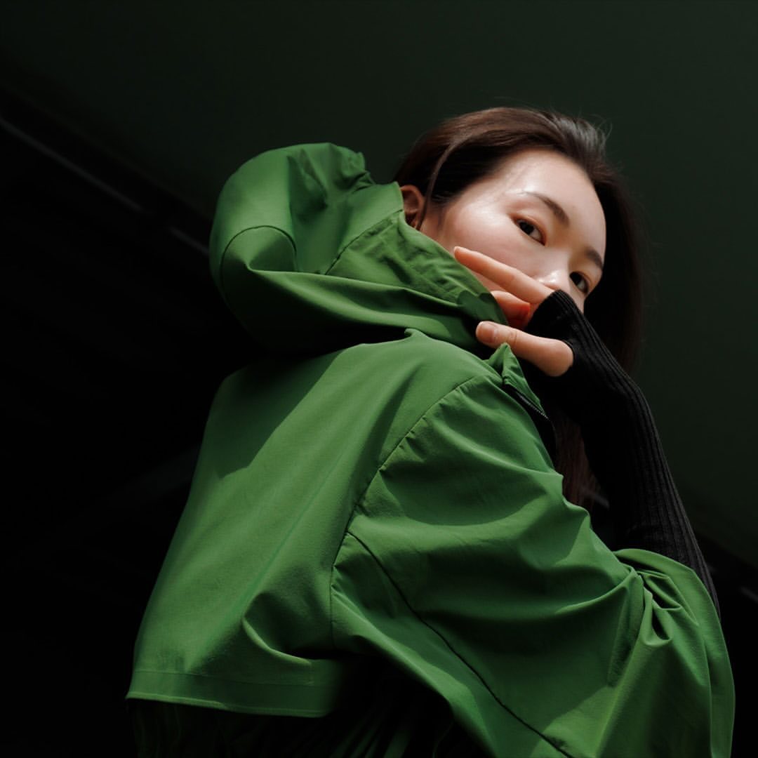 Adult woman in a green hooded jacket with black sleeves poses indoors, obscuring part of her face with her hand.