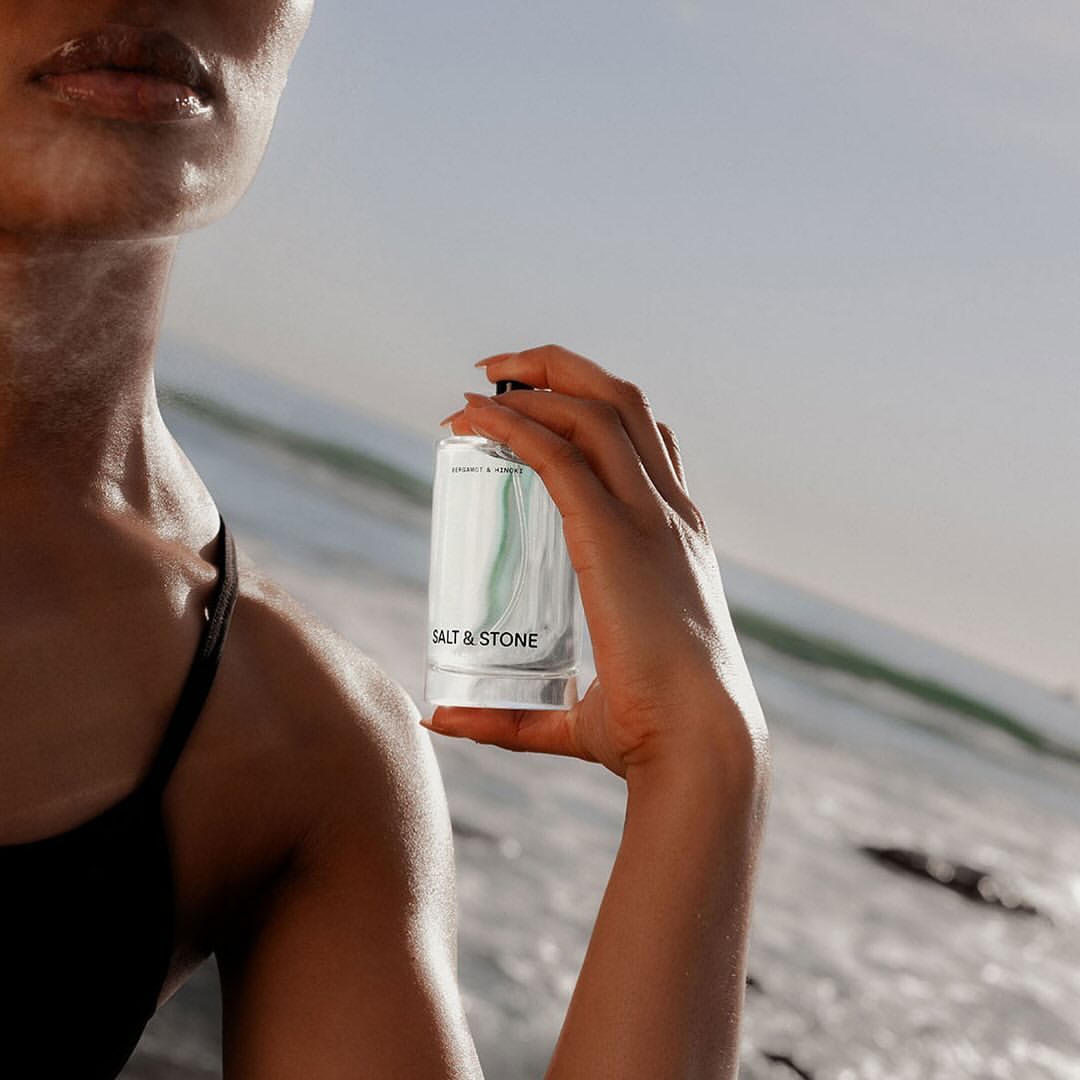 Adult female holds a bottle of Salt & Stone lotion in front of a white sand dune and a clear sky, in a product shot.