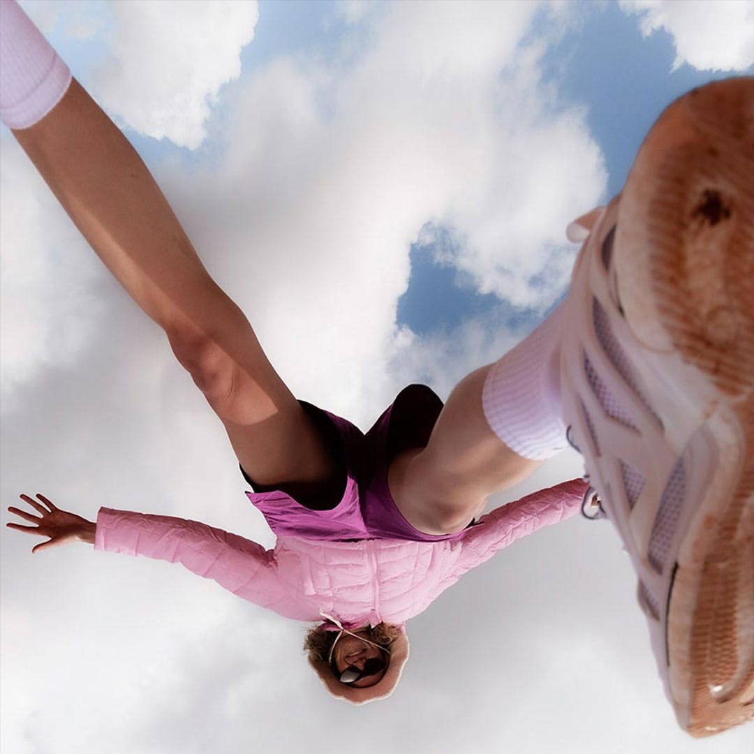A woman in athletic wear is viewed from a low angle against a sky with clouds, showcasing her running shoes, arms outstretched, and pink jacket.