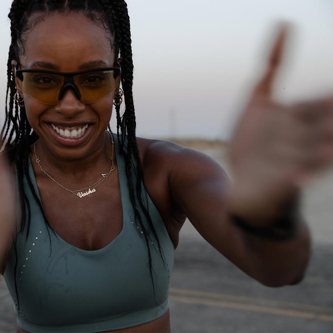 Smiling African-American woman in a sports bra and sunglasses, posing with arm extended on a paved surface with a blurred hand.