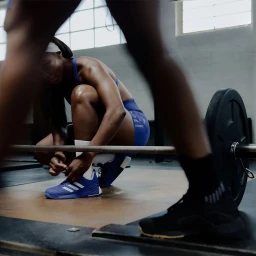 Athletic woman in blue sports bra and shorts kneeling to tie her blue sneakers in a gym with weights.