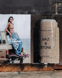 Wheatpaste posters on a weathered wall featuring a woman in a bikini and jeans leaning on a beige SUV and a deodorant product package