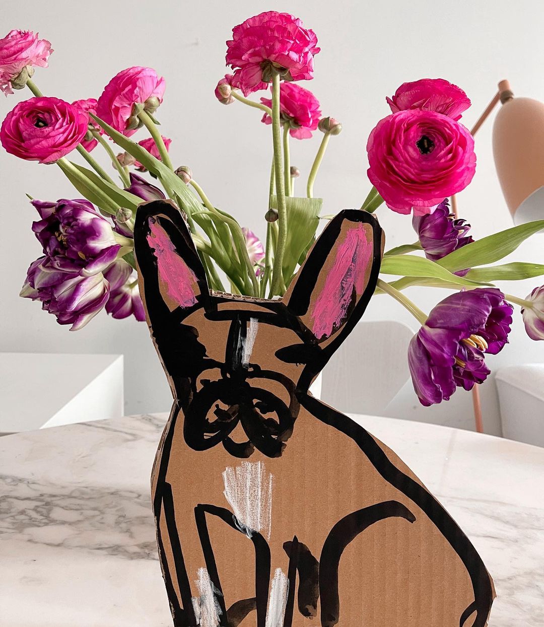 Ranunculus and tulip floral arrangement with cardboard dog vase on a marble table against a bright white background in a studio.