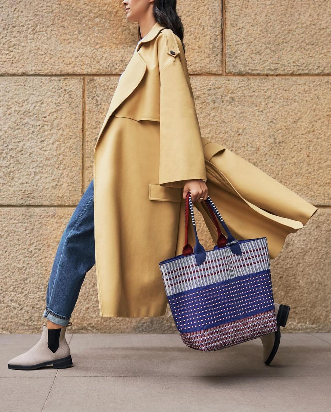 Woman in a trench coat walks on the sidewalk, carrying a patterned tote bag, wearing jeans and boots. Neutral background of textured stone.