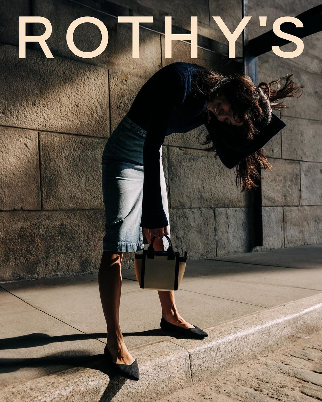 A woman in a skirt and blouse holding a purse and looking down, stands on a city street, with the brand Rothy's logo above her.