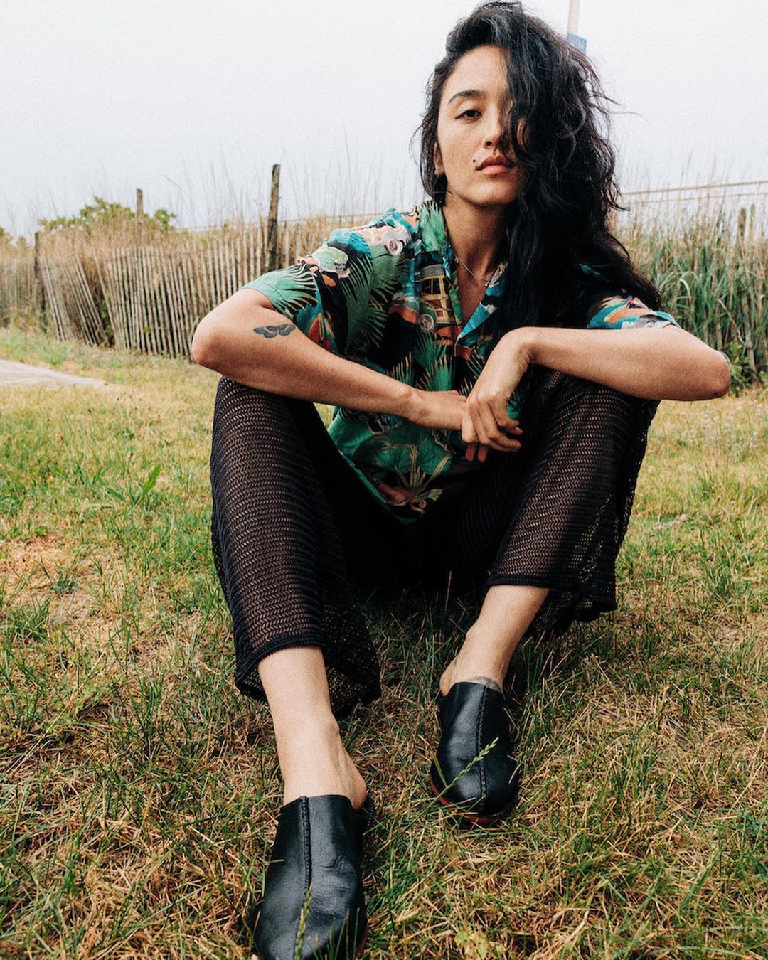 A woman with long dark hair in a patterned shirt and black pants sits cross-legged on a lawn against a background of fences and foliage.