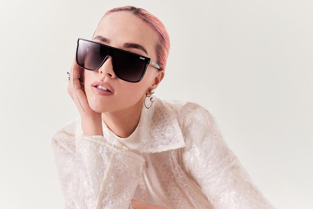 Close-up of a fair-skinned person with pink hair and large black sunglasses, posing indoors against a pale backdrop in a textured white top.