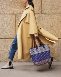 Woman walking outdoors wearing a beige trench coat, blue jeans, and carrying a patterned blue tote bag.