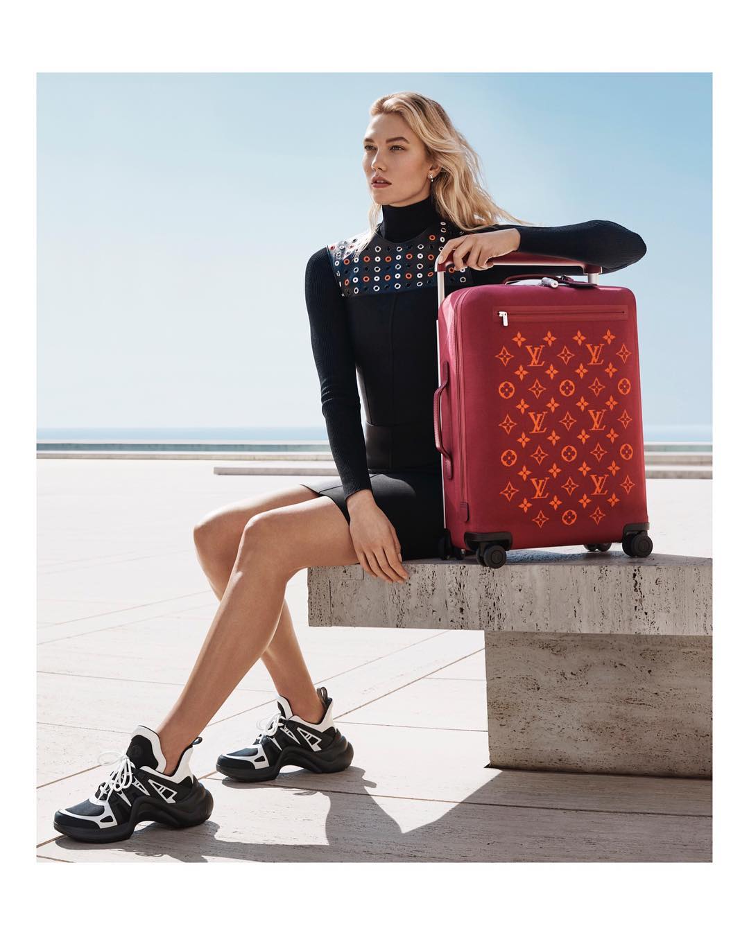 Woman poses with Louis Vuitton suitcase outdoors, wearing black dress and sneakers, with ocean and sky in the background. Fashion photography.