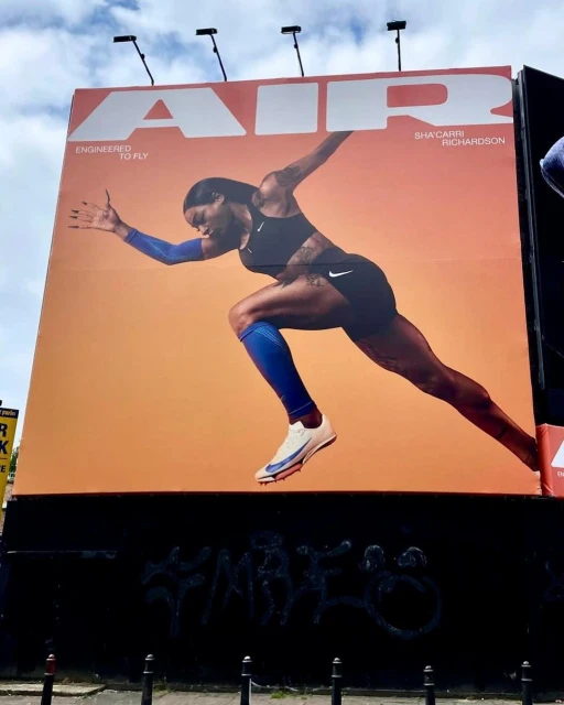 Sha'Carri Richardson featured on a Nike billboard in running pose wearing a sports bra and shorts against an orange background.