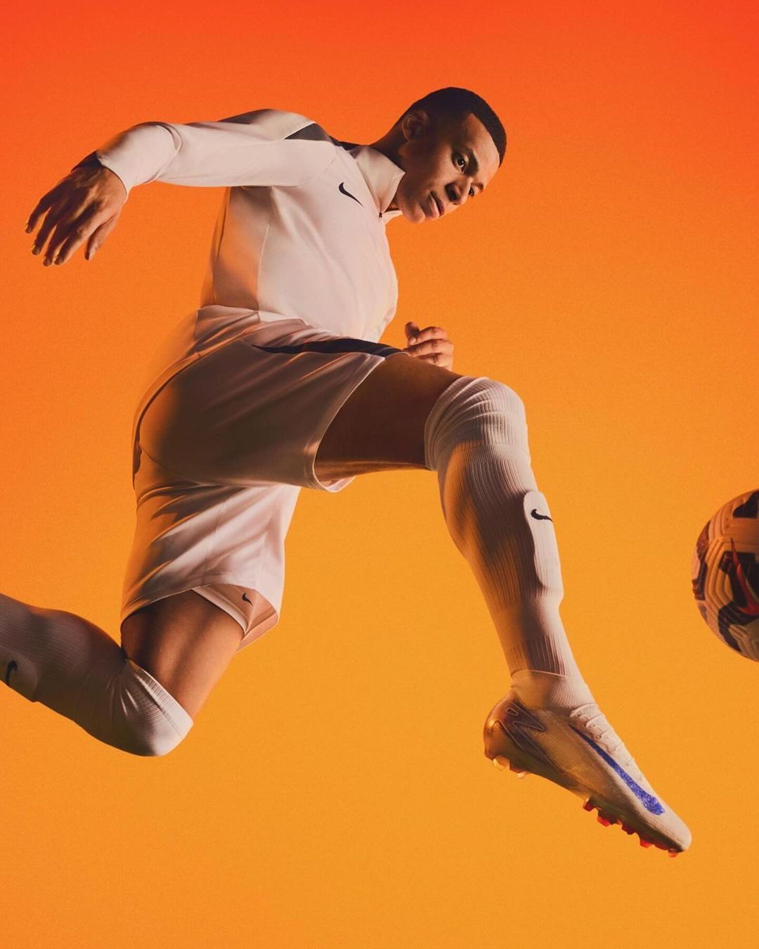 Male soccer player mid-air, kicking a soccer ball against a gradient orange background, wearing white athletic apparel and footwear.
