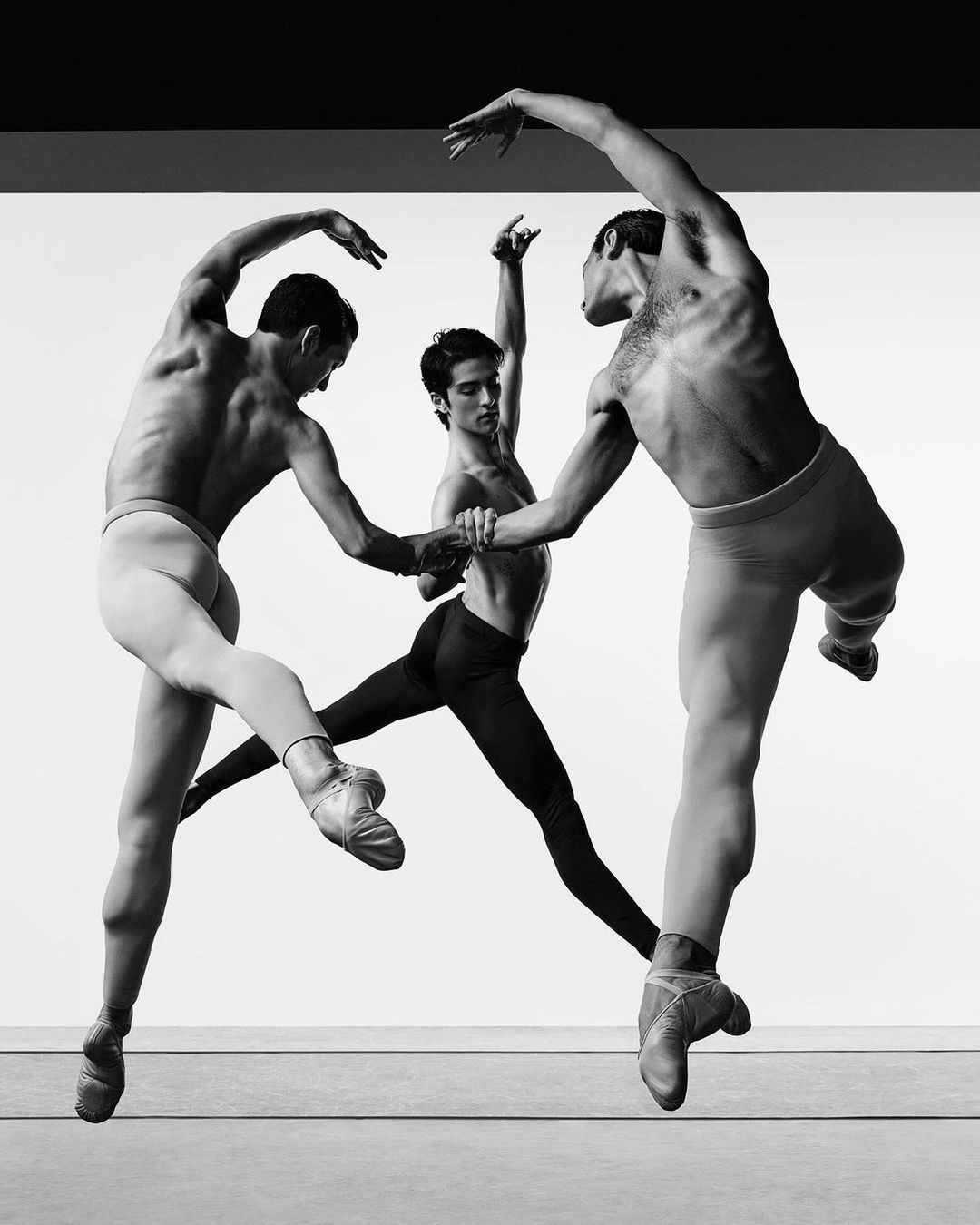 Three male ballet dancers leap and pose in a black and white studio photograph against a white backdrop, showcasing athletic skill.