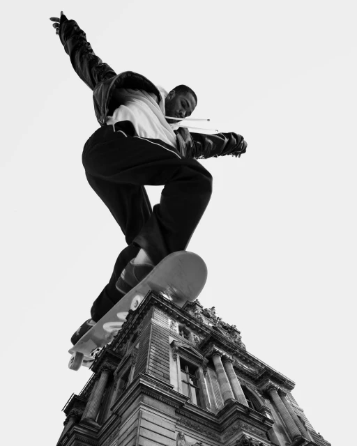Black and white photo of an African American male skateboarder mid-air over an ornate building, captured from a low angle, emphasizing motion and architecture.