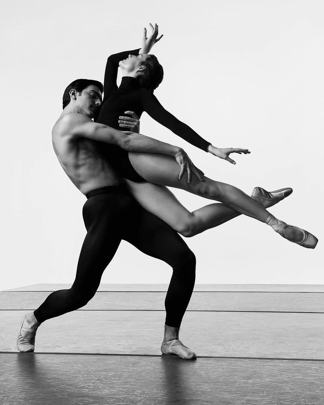 Black and white image of ballet dancers, a male dancer holding a female dancer in a lift pose against a plain background. 