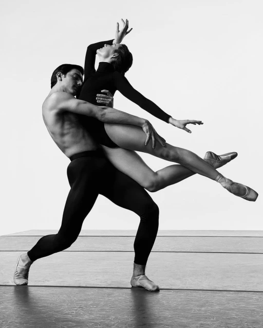 Black and white image of ballet dancers, a male dancer holding a female dancer in a lift pose against a plain background. 