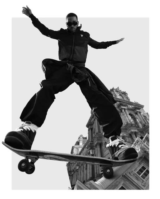 Black and white image of a man skateboarding in front of a building. The man is wearing sunglasses and has his arms outstretched.