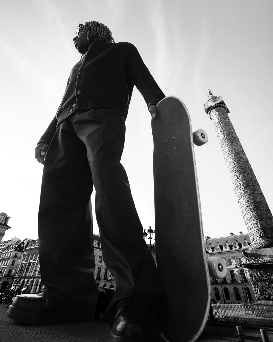 A low angle shot of a man with dreadlocks holding a skateboard in a city square with historical buildings and a tall column in the background.