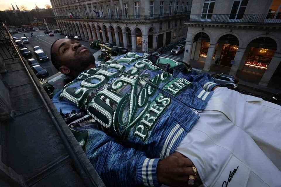 Adult male poses in an embellished jacket and white pants on a rooftop, set against a cityscape with buildings and street traffic.