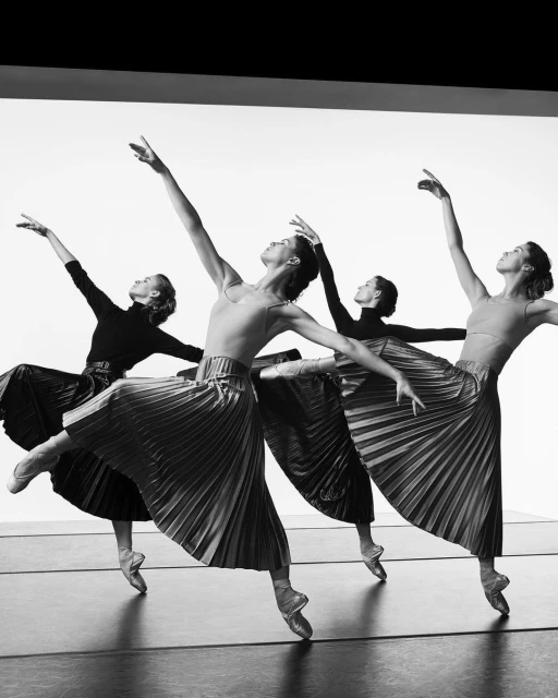 Four ballet dancers in black and gray skirts and leotards in pointe shoes, arms raised in studio, black and white stage photography.