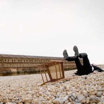A man in a black suit lies on a pebble beach with an overturned chair next to him, under an overcast sky near a wall.