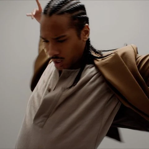 Man with braided hair wearing a tan shirt and brown jacket posing in front of a plain grey background in a studio setting.