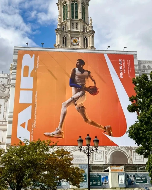 Large outdoor advertisement of basketball player Victor Wembanyama in athletic wear holding a basketball in front of a European building.