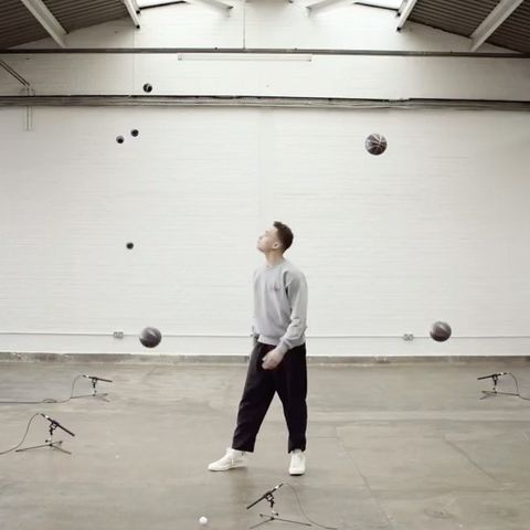 Male juggler in gray sweatshirt with basketballs in bright white room. Multiple microphones also visible on the concrete floor.