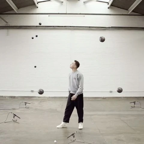 Male juggler in gray sweatshirt with basketballs in bright white room. Multiple microphones also visible on the concrete floor.