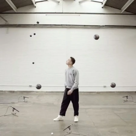 Male juggler in gray sweatshirt with basketballs in bright white room. Multiple microphones also visible on the concrete floor.