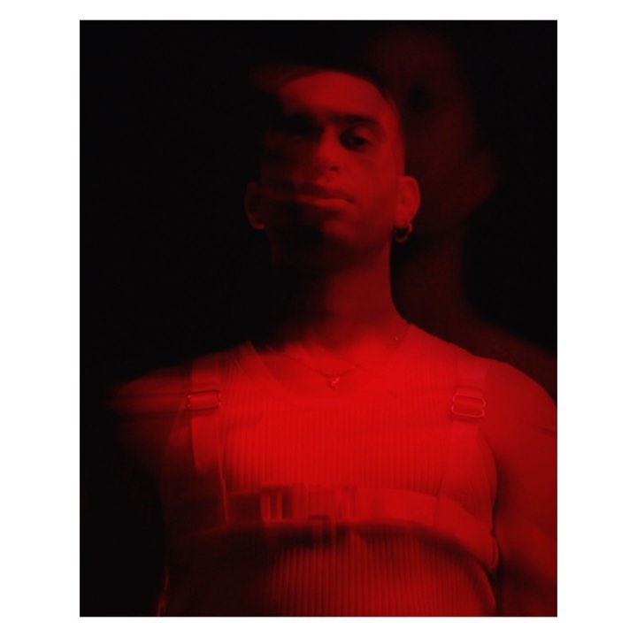 Portrait of a man with dark hair in red lighting, wearing a white ribbed shirt and harness, standing against a black background.