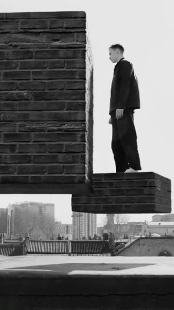 Monochrome shot of a man in dark clothing standing on a brick platform with a city skyline in the background on a bright, sunny day.