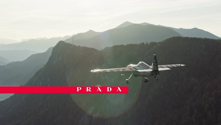 An airplane flies above mountain ranges and below a title card with the word PRADA in all caps, highlighting aviation and nature.