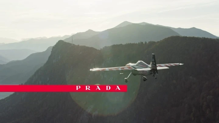 An airplane flies above mountain ranges and below a title card with the word PRADA in all caps, highlighting aviation and nature.