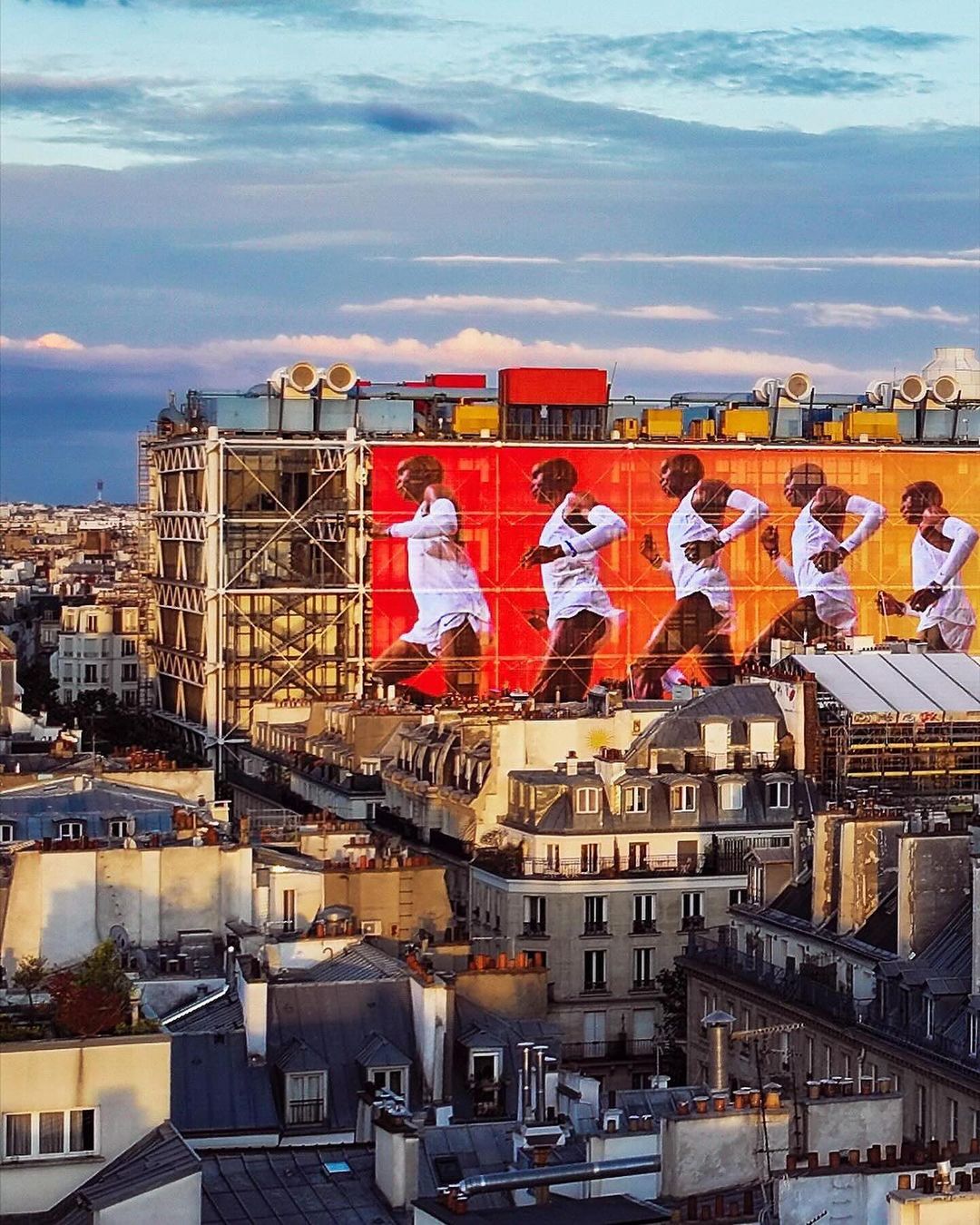 Multiple exposures show a Black male figure running across the top of an orange building with the Paris cityscape visible in the background.