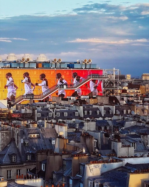 Cityscape view of Paris rooftops with rooftop art installation featuring silhouette figures on a building with an orange and yellow backdrop.