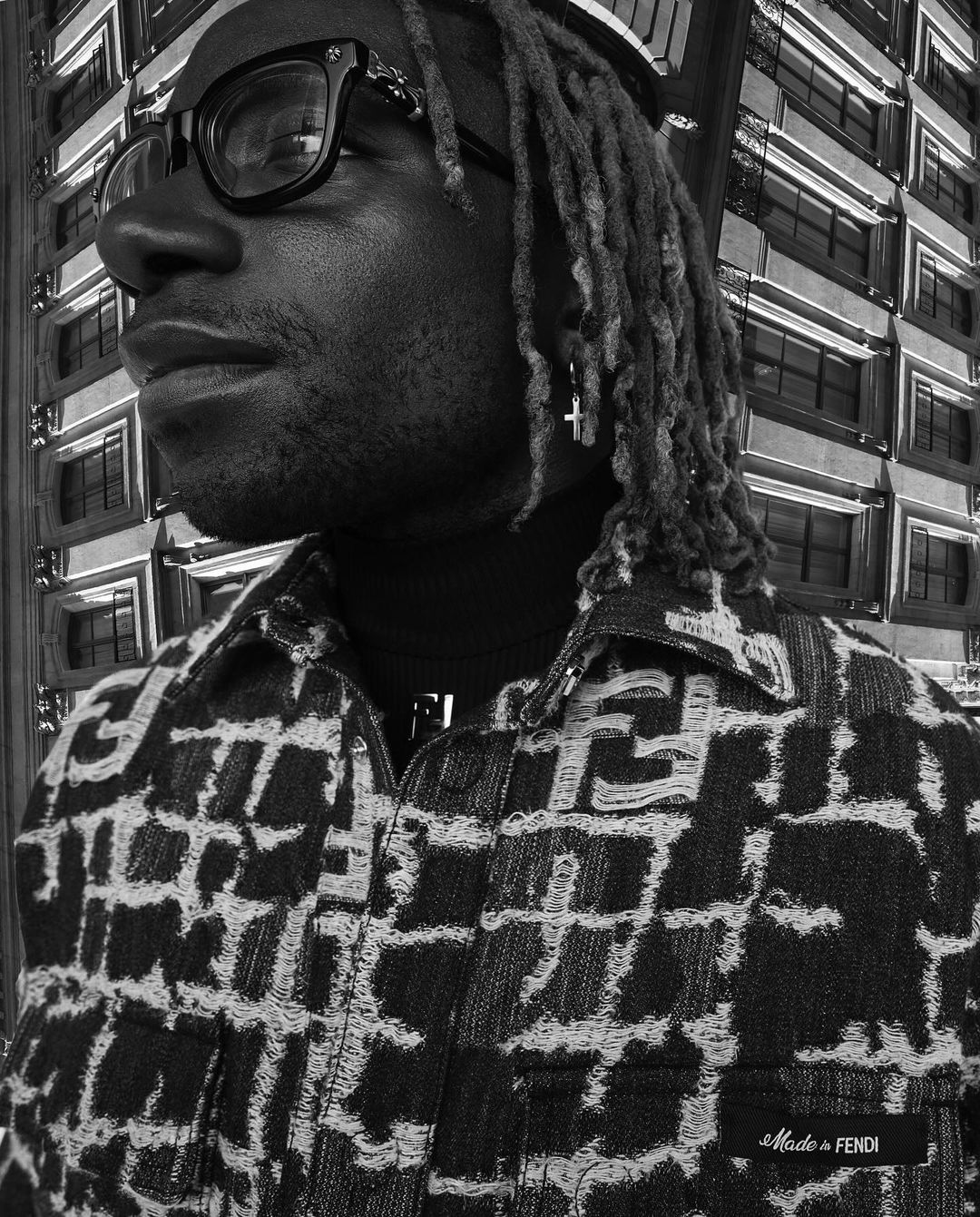 Monochrome portrait of a stylish Black male with locs, glasses, and a patterned jacket standing against a high-rise building backdrop.