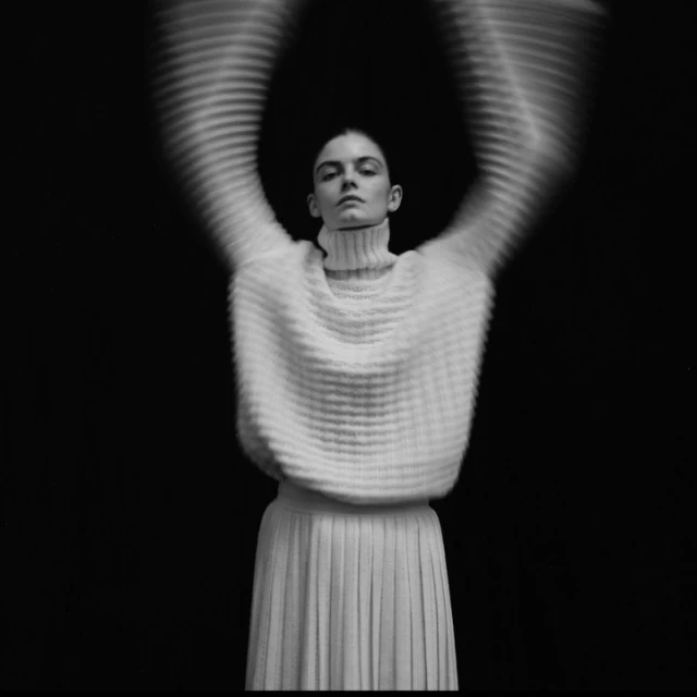 Monochrome portrait of a woman in a turtleneck sweater and pleated skirt with blurred arm motion against a black background.
