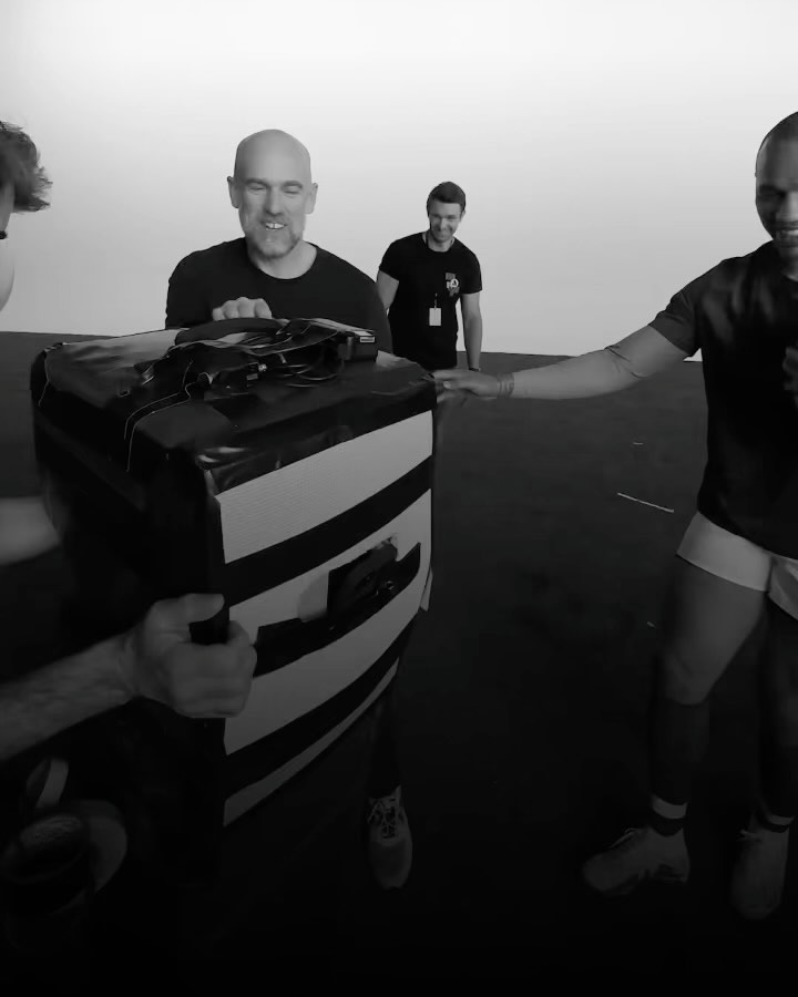 Four adult male rugby players are practicing a drill with a pad in an indoor training facility, with black flooring and a white background.
