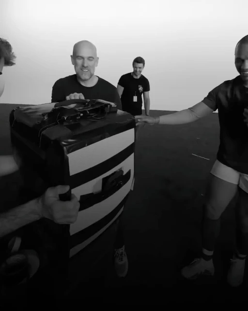 Four adult male rugby players are practicing a drill with a pad in an indoor training facility, with black flooring and a white background.