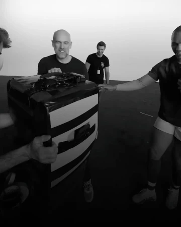 Four adult male rugby players are practicing a drill with a pad in an indoor training facility, with black flooring and a white background.