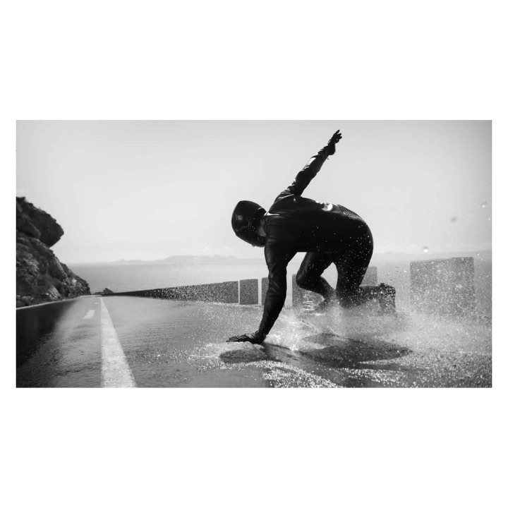 Monochrome shot of a surfer in a wetsuit dynamically riding the water on a paved road, with coastal cliffs and the ocean in the distance.