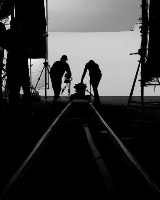 Monochrome film production scene: two crew members prepare a camera dolly as an actor runs in the background on an outdoor set.