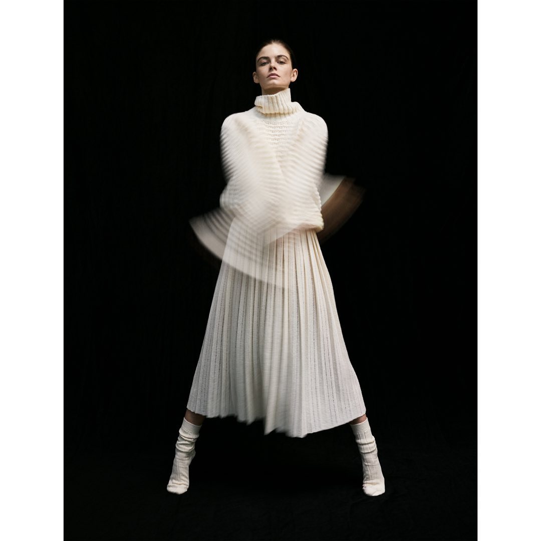 Elegant woman in flowing white turtleneck sweater and pleated skirt strikes a pose against a stark black backdrop, motion blur on arms.