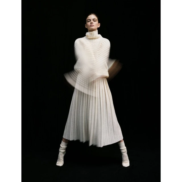 Elegant woman in flowing white turtleneck sweater and pleated skirt strikes a pose against a stark black backdrop, motion blur on arms.